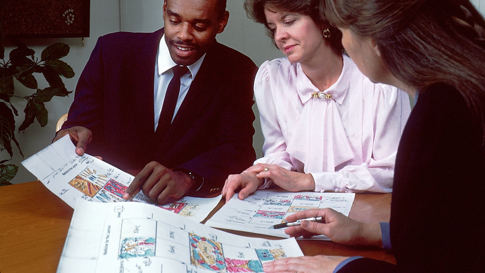Three people at a conference table discussing illustrated storyboards, used as a visual analogue for multiple minds examining one problem