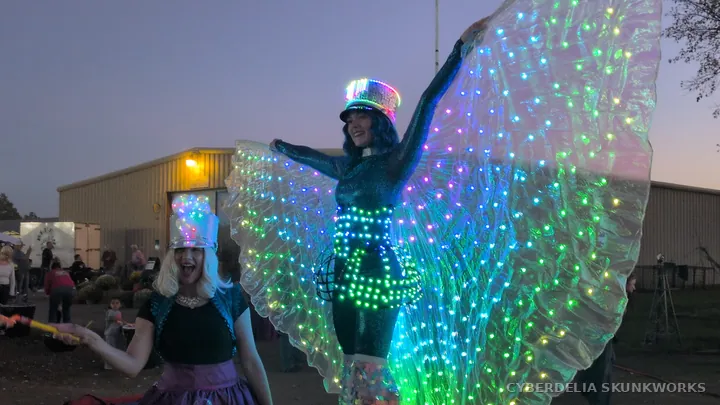 An Inspyral Circus performer raises luminous wings beside the midway.